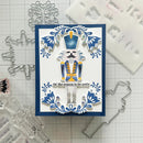 A handmade Christmas nutcracker card featuring a layered paper nutcracker with blue, white, and gold details, surrounded by blue floral scrollwork. The card is displayed on a gridded craft mat with metal and clear stamping tools around it.