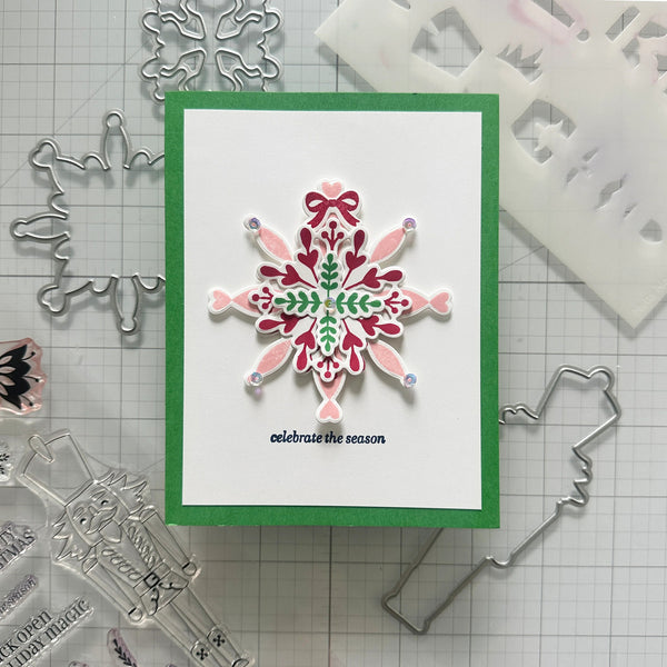 A handmade holiday greeting card featuring a layered paper snowflake design in pink, green, and burgundy on a white background with a green border. The card is displayed on a gridded cutting mat surrounded by metal and clear stamping/cutting dies.