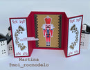 A handmade holiday pop-up card featuring a nutcracker illustration in the center, flanked by white panels with gold floral designs and red borders. The left panel includes the text 'crack open the holiday magic' and the name Martina with handle @moi_rocnodeleo is printed below the card.