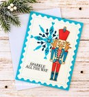 A handmade holiday greeting card featuring a nutcracker and a blue snowflake design with the text 'SPARKLE ALL THE WAY'. The card is styled on a light wood background with a white envelope and pine branches.