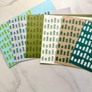 Six patterned paper sheets or envelopes fanned out on a light marble surface. Each sheet has a repeating pine tree motif in a different base color: light blue, pale lavender, olive green, white, kraft beige, and dark teal.