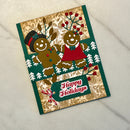 A handmade Christmas greeting card featuring two gingerbread men, one in a top hat and the other in a red dress, with a 'Happy Holidays' banner. The card is decorated with green borders, a gold and beige floral patterned background, and small white pine trees, photographed on a light textured fabric surface.