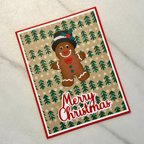 A handmade Christmas greeting card featuring a gingerbread man with a top hat on a patterned background of green pine trees, with 'Merry Christmas' written in red and white script. The card is photographed on a light gray textured fabric background.