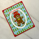 A handmade Christmas greeting card featuring a gingerbread man illustration, a candy cane, and the text 'Merry Christmas' on a light blue background with green pine tree patterns and a red border, photographed on a light marble surface.