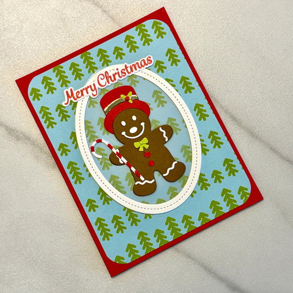 A handmade Christmas greeting card featuring a gingerbread man illustration, a candy cane, and the text 'Merry Christmas' on a light blue background with green pine tree patterns and a red border, photographed on a light marble surface.