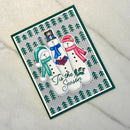 A handmade holiday greeting card featuring three snowmen wearing colorful winter hats and scarves, with the phrase 'Tis the Season' in script. The card has a gray background with a border of green pine trees and is photographed on a light beige textured fabric surface.