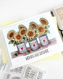 A handmade greeting card featuring three potted sunflowers with the text 'WISHING YOU SUNSHINE' at the bottom, styled on a yellow cardstock with stamping tools nearby.