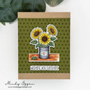 A handmade greeting card with a kraft paper border and an olive green polka dot center, featuring a die-cut illustration of three sunflowers in a tin can, with the text 'WISHING YOU SUNSHINE' below. The card is displayed on a white surface with green leaves in the background, and the brand name 'Mindy Eggen' is visible in the bottom left corner.
