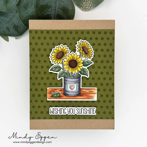 A handmade greeting card with a kraft paper border and an olive green polka dot center, featuring a die-cut illustration of three sunflowers in a tin can, with the text 'WISHING YOU SUNSHINE' below. The card is displayed on a white surface with green leaves in the background, and the brand name 'Mindy Eggen' is visible in the bottom left corner.
