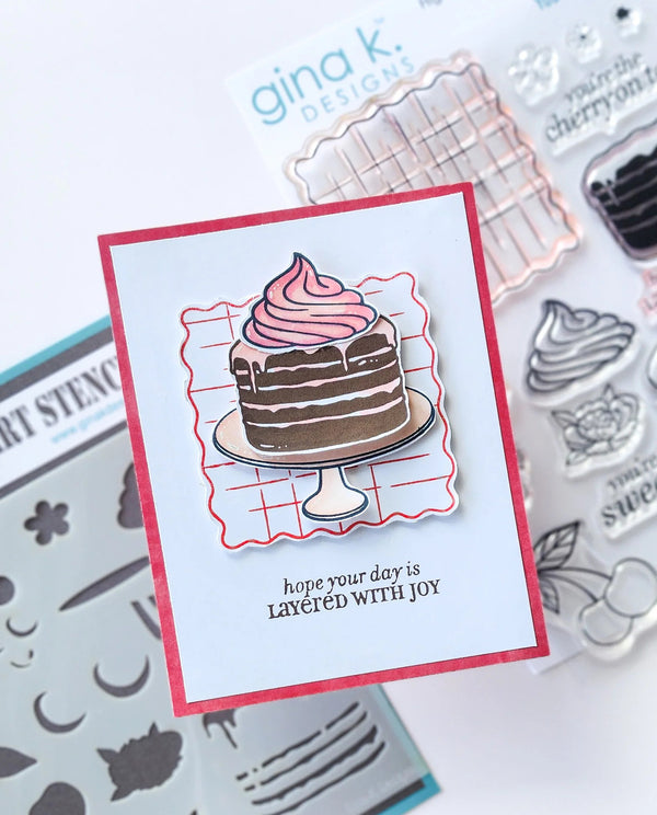 A handmade layered cake card with a pink frosting top and the printed message 'hope your day is LAYED WITH JOY', displayed on top of Gina K. Designs stamp and stencil kits.