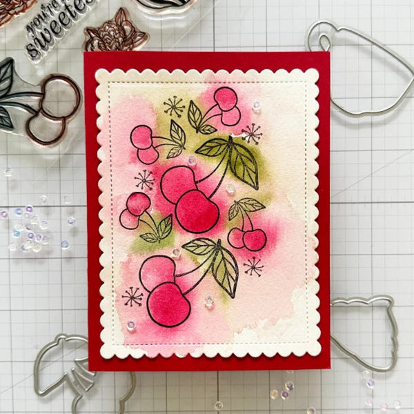 A handmade greeting card featuring a cherry illustration, displayed on a gridded craft mat with metal cutters and clear stamps around it.