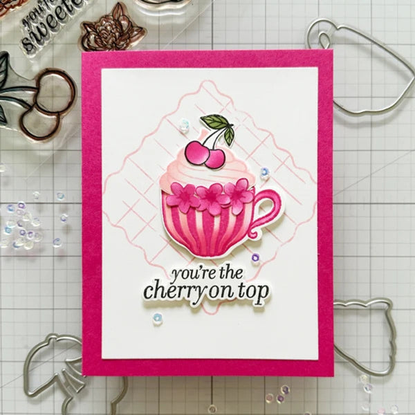 A handmade greeting card with a bright pink border and a white center panel, featuring an illustrated pink striped teacup with cherries and the phrase 'you're the cherry on top.' The card is photographed on a gridded craft mat with metal dies and embellishments around it.