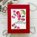 A handmade greeting card with a bright red background and a white central panel featuring cherry illustrations and the phrase 'you're the cherry on top.' The card is photographed on a white grid mat with craft tools and embellishments around it.
