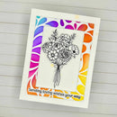 A handmade greeting card featuring a black-and-white floral bouquet illustration in the center, surrounded by a colorful patterned border in pink, purple, blue, orange, and yellow. The card reads 'Sending loving wishes your way' and is photographed on a white wood plank background.