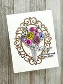 A handmade greeting card featuring a glittery gold ornate frame, a colorful floral bouquet with yellow, pink, purple, and blue flowers, and the words 'Need you more than you'll ever know' on a white card, displayed on a rustic gray wood background.