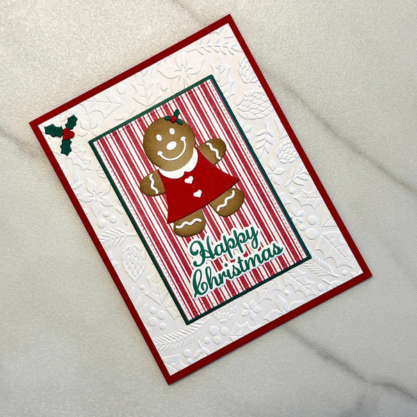A handmade Christmas greeting card featuring a gingerbread man in a red dress, a red and green striped central panel, and the words 'Happy Christmas' in green script. The card has a red border and a white embossed background with subtle floral and snowflake patterns, photographed on a light textured surface.