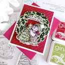 A handmade Christmas card featuring a penguin sitting on a small stool beside a decorated tree, with the text 'Deck the Halls with Lots of Joy!'. The card is layered over a red textured background and pink envelope, with Gina K.