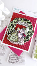 A handmade Christmas greeting card featuring a penguin wearing a green hat and red scarf, sitting on a stool next to a decorated Christmas tree. The card has a red quilted patterned background, a green wreath with black holly accents, and the text 'Deck the Halls with Lots of Joy!'.
