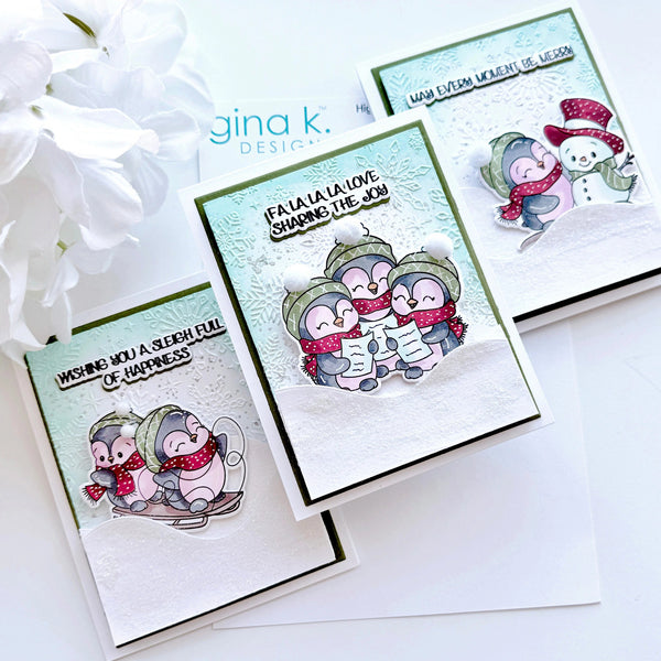 Three handmade holiday greeting cards with penguin illustrations, arranged on a white surface with white flowers in the corner. The cards feature layered white and mint green backgrounds, black borders, and text including 'Wishing you a sleigh full of happiness' and 'Fa la la la love sharing the joy'.