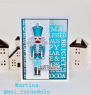 A handmade Christmas greeting card featuring a die-cut nutcracker illustration, set against a white background with decorative miniature houses on either side. The card has a turquoise and white color scheme with festive text.