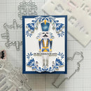 A handmade Christmas nutcracker card featuring a die-cut nutcracker figure on a white and navy blue layered background, surrounded by crafting tools and metal cutters on a gridded surface.