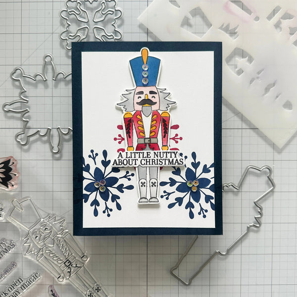 A handmade Christmas nutcracker card featuring a colorful stamped nutcracker figure with the text 'A LITTLE NUTTY ABOUT CHRISTMAS' and blue floral motifs, displayed on a gridded cutting mat with metal and plastic craft dies around it.