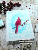 A handmade greeting card featuring a red cardinal perched on a branch with red berries, set against a light blue circular background and a white card with festive text. The card is styled on a rustic wooden surface with holiday decorations.