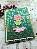 A handmade Christmas greeting card with a green background, gold glitter holiday text, a pink bow, and a green ornament cutout, photographed on a rustic white wood surface with red berries as props.