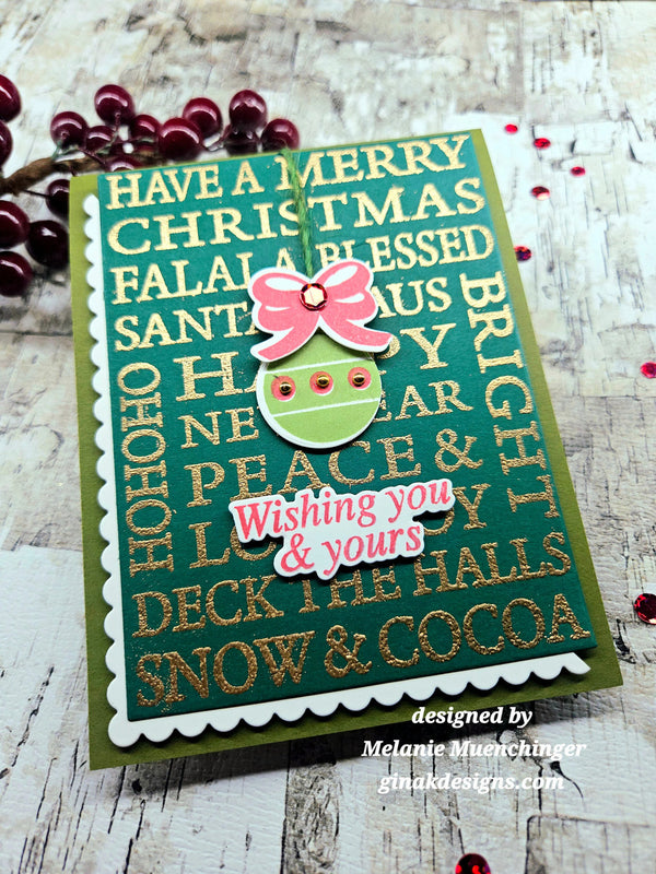 A handmade Christmas greeting card with a green background, gold glitter holiday text, a pink bow, and a green ornament cutout, photographed on a rustic white wood surface with red berries as props.