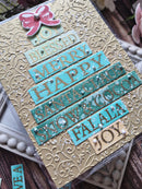 A handmade Christmas greeting card featuring a gold embossed background with layered turquoise and gold text strips reading 'BLESSED', 'MERRY', 'HAPPY', 'SANTA', 'GLORY', 'SNOW', 'AND COCO', 'FALALA', and 'JOY'. A pink bow embellishment sits at the top left.