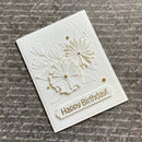 A handmade white card with embossed floral details, small gold dots, and a banner reading 'Happy Birthday!' in gold lettering, photographed on a gray script-patterned background.