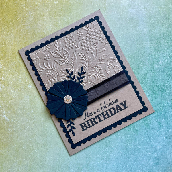 A handmade greeting card with a beige base, featuring a navy blue scalloped border, an embossed floral pattern, a matching navy blue paper flower with a crystal center, and the text 'Have a fabulous BIRTHDAY'. The card is photographed at an angle on a textured, iridescent yellow-to-blue background.
