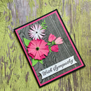 A handmade sympathy greeting card featuring layered pink and white paper flowers on a dark wood-grain background, bordered with pink and black, and a white banner reading 'With Sympathy' at the bottom, photographed on a distressed green wood surface.