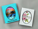 Two handmade greeting cards displayed on a neutral gray background. The card on the left is bright turquoise with a textured border and a circular cutout showing a tropical sunset scene with palm trees.