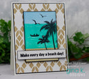 A handmade greeting card featuring a tropical beach scene with palm trees, a small boat, and the text 'Make every day a beach day!'. The card has a white and gold patterned border, a turquoise ocean scene, and black silhouettes, photographed against a blurred green plant background.
