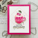 A handmade greeting card with a bright pink border and a white center panel, featuring an illustrated pink striped teacup with cherries and the phrase 'you're the cherry on top.' The card is photographed on a gridded craft mat with metal dies and embellishments around it.