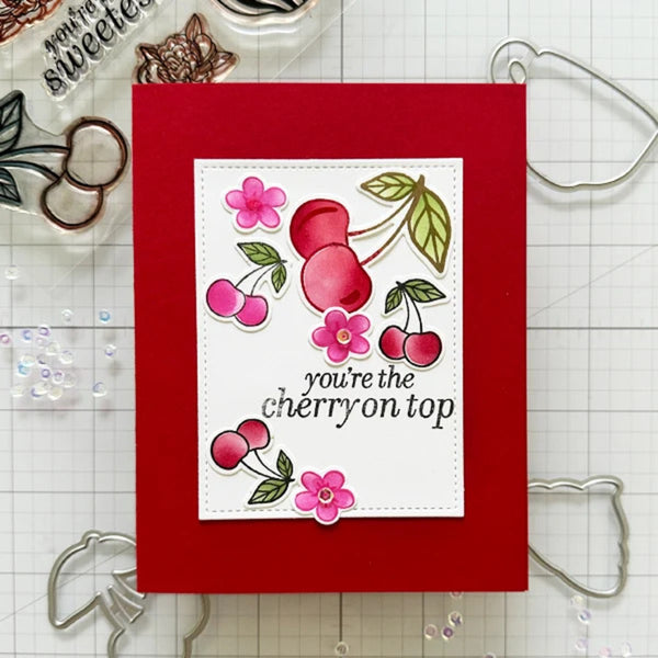 A handmade greeting card with a bright red background and a white central panel featuring cherry illustrations and the phrase 'you're the cherry on top.' The card is photographed on a white grid mat with craft tools and embellishments around it.