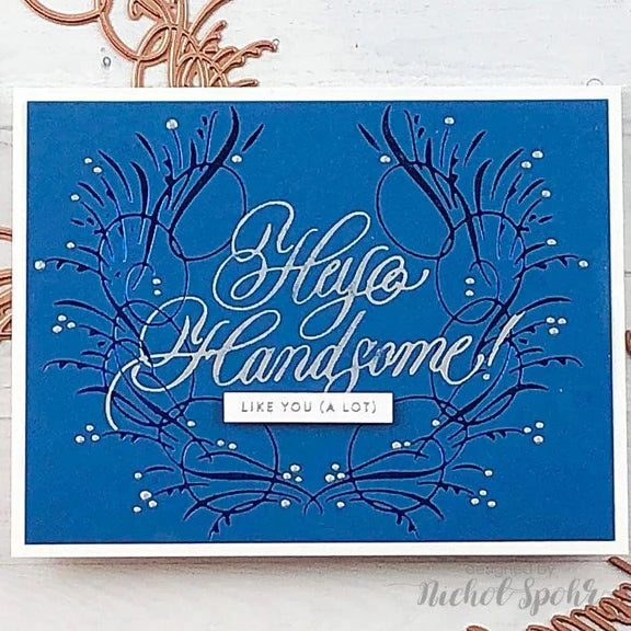 A square blue greeting card with white and dark blue decorative script and floral line art, plus the phrase 'Hello Handsome! LIKE YOU (A LOT)' in the center, photographed on a light surface with copper-colored decorative elements around it.