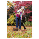 A woman in a colorful knit sweater and dark blue wide-leg jeans rakes fallen leaves with a black-handled rake in an autumn garden. A wire basket filled with leaves sits on the grass nearby.