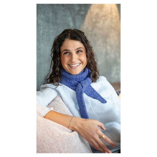 A smiling woman with curly brown hair wearing a textured blue knit scarf and a white top, seated indoors with a neutral gray and beige background.
