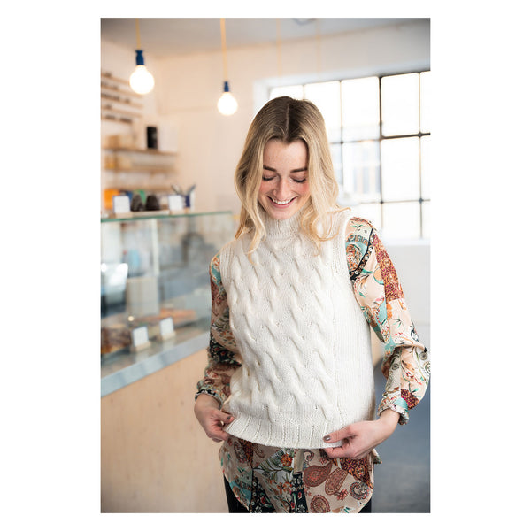 A woman smiling in a cozy indoor shop setting, wearing a cream cable-knit sweater vest over a long-sleeve paisley blouse.