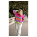 A woman wearing a vibrant, multicolored striped knit sweater and white pants walking outdoors on a paved path with trees in the background.