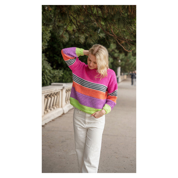 A woman wearing a vibrant, multicolored striped knit sweater and white pants walking outdoors on a paved path with trees in the background.