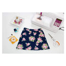 A navy blue floral skirt with pink roses and green leaves is laid flat on a white surface, surrounded by a white sewing machine, scissors, thread, and a small dish of decorative items.