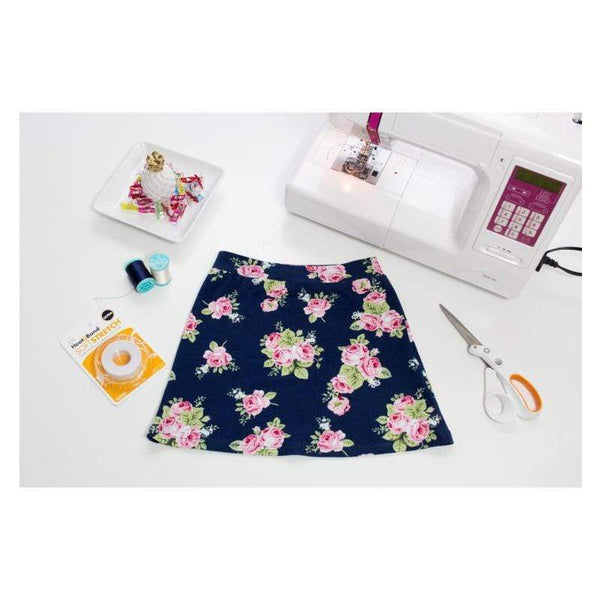 A navy blue floral skirt with pink roses and green leaves is laid flat on a white surface, surrounded by a white sewing machine, scissors, thread, and a small dish of decorative items.