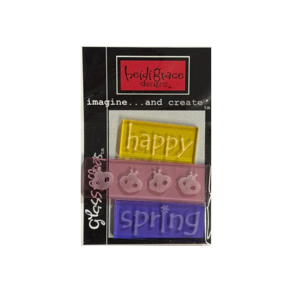 A packaged set of three embossed acrylic tiles  featuring the words 'happy', 'spring',  in yellow, pink, and purple, respectively, on a black backing card.