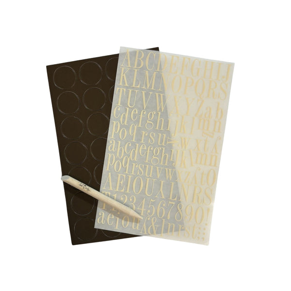 A sheet of cream alphabet and number decals on a translucent backing, placed over a dark brown circular sticker sheet, with a wooden spatula tool resting diagonally on a white background.