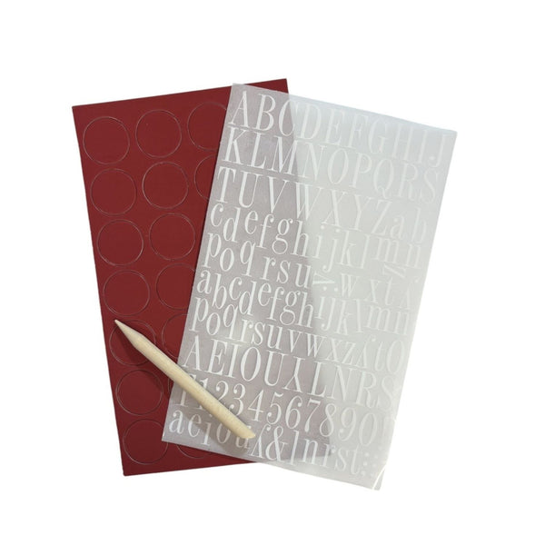 A sheet of clear alphabet and number stickers overlaid on a dark red sheet of circular stickers, with a wooden stick tool placed diagonally across the front, all on a plain white background.