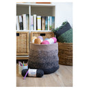 A large round crocheted storage basket in a heathered gray and black colorway sits on a light wood floor in a styled craft room setting, surrounded by other yarn skeins, a cup of knitting needles, and woven baskets against a white bookshelf.