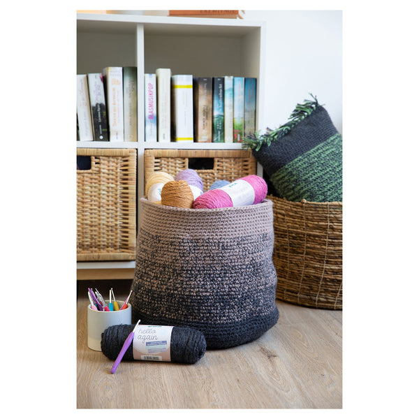 A large round crocheted storage basket in a heathered gray and black colorway sits on a light wood floor in a styled craft room setting, surrounded by other yarn skeins, a cup of knitting needles, and woven baskets against a white bookshelf.